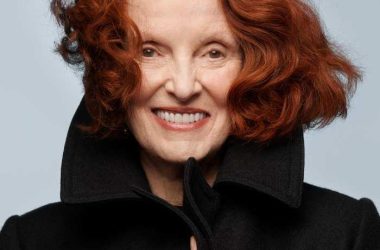 Grace Coddington with her signature red hair wearing a black coat in a studio portrait.