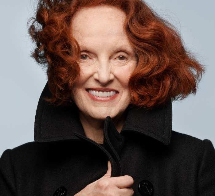 Grace Coddington with her signature red hair wearing a black coat in a studio portrait.
